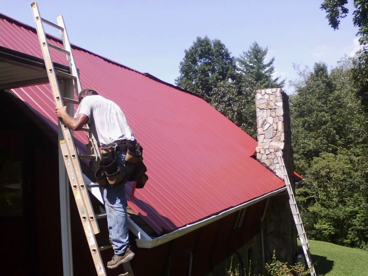 Roofer on a ladder beside a red metal roof and stone chimney — Soffit & Fascia Repair by Roofing Brothers in Marshfield
